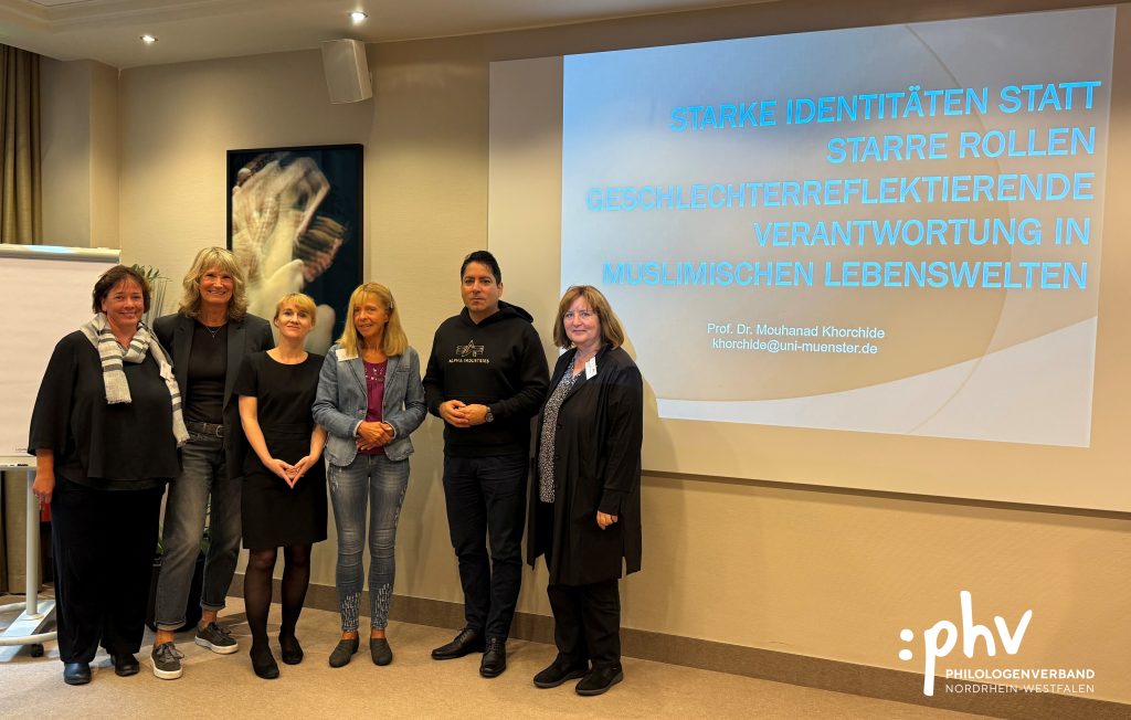 Eine Gruppe von sechs Personen steht nebeneinander in einem Seminarraum und lächelt in die Kamera. Rechts im Bild ist eine große Leinwand mit der Präsentationsfolie „Starke Identitäten statt starrer Rollen – Geschlechterreflektierende Verantwortung in muslimischen Lebenswelten“ von Prof. Dr. Mouhanad Khorchide, Universität Münster. Links steht ein Flipchart, rechts unten ist das Logo des Philologenverbands Nordrhein-Westfalen zu sehen.