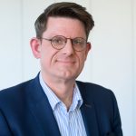 Brustbild eines Mann mit dunklem Sakko und Brille, der nett in die Kamera lächelt.