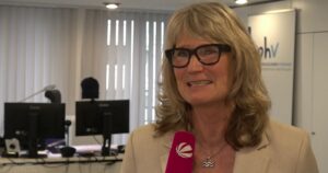 Eine Frau mit langen blonden Haaren und schwarzer Brille spricht in ein Mikrofon.